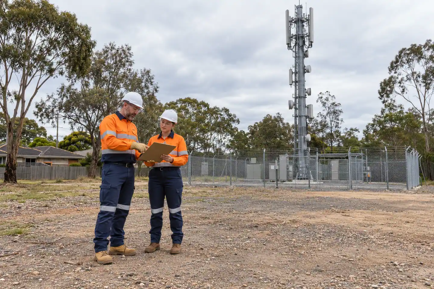 Two supervisors review a clipboard outside a fenced telecommunications-tower enclosure.