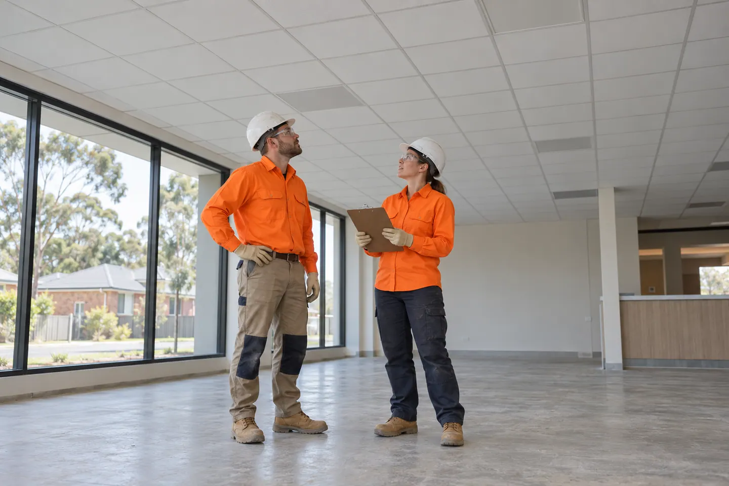 Two shopfitters inspect a suspended ceiling from ground level.